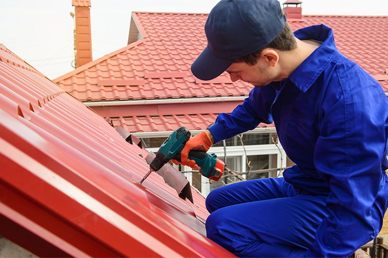 Young man worker in blue overall fix a metal tile roof with screwdriver. Roofing work concept
